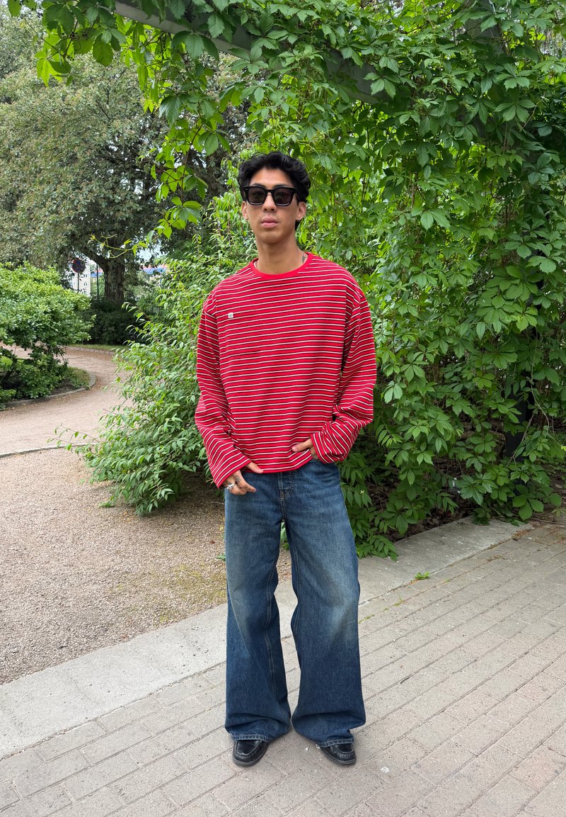 Jeune homme portant des lunettes de soleil noires, une chemise à manches longues rayée rouge et blanche, un jean large bleu, debout sur un chemin avec des arbres verts en arrière-plan.