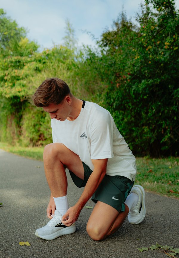 Camiseta deportiva blanca con detalles y logo en negro, pantalones cortos verdes, zapatillas blancas con detalles en negro, atándose los cordones en un camino rodeado de vegetación.