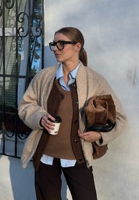 Μπεζ χνουδωτό παλτό, καφέ πλεκτό γιλέκο πάνω από μπλε πουκάμισο, σκούρο παντελόνι με ρίγες, κρατώντας ένα φλιτζάνι καφέ και μια καφέ τσάντα με μαύρη ζώνη.