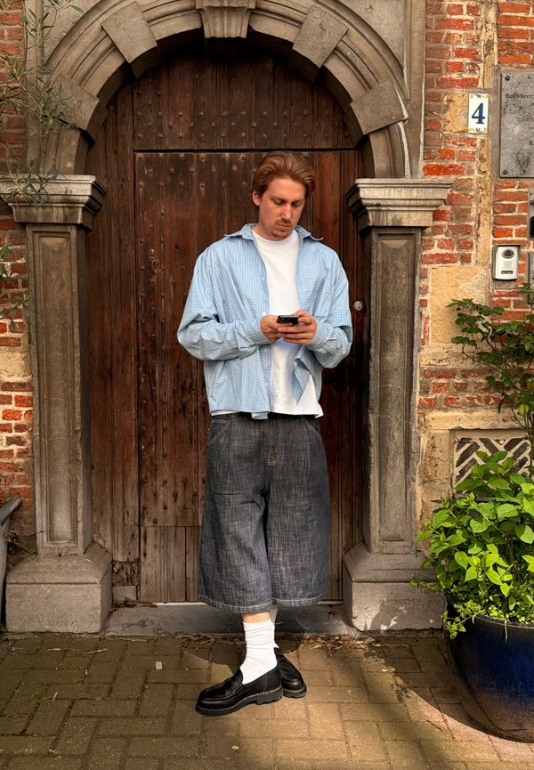 Man in blauw geruit overhemd en zwarte korte broek staand op een bakstenen pad, kijkend naar zijn telefoon voor een grote houten boogdeur.