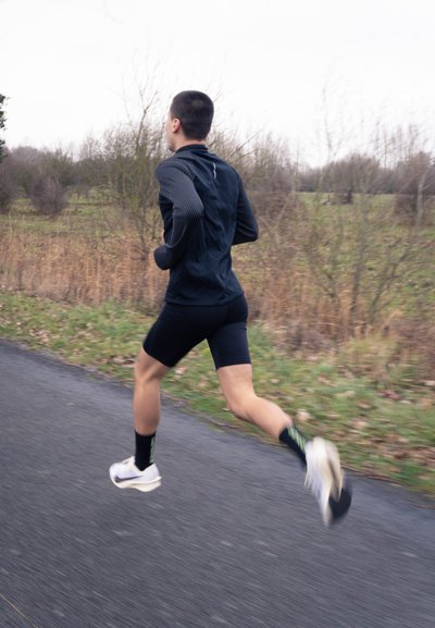 Hombre joven con ropa deportiva negra corriendo por un camino pavimentado a través de un paisaje rural cubierto de hojas, con árboles desnudos.