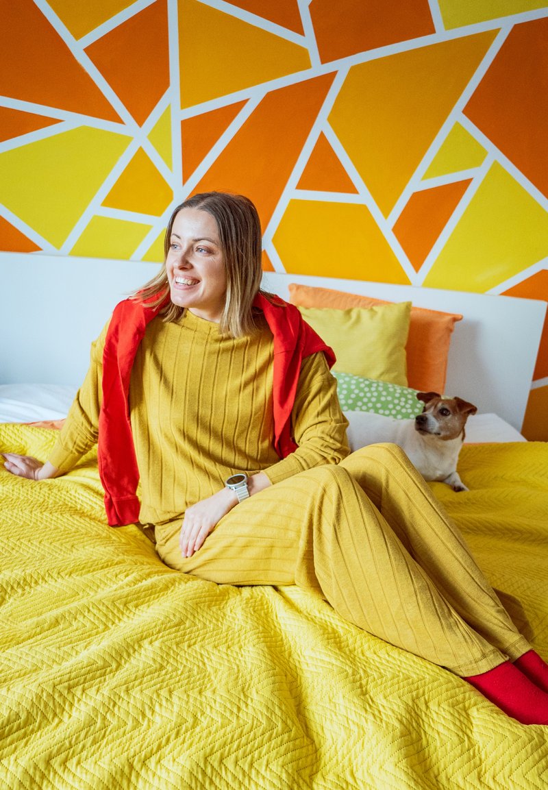 Femme en tenue jaune et chaussettes rouges assise sur une couverture de lit jaune avec un petit chien à ses côtés, devant un mur géométrique lumineux orange et jaune.