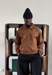 Homme portant un bonnet noir et une veste marron, debout avec les mains dans les poches devant une étagère en bois contenant des livres et des objets.