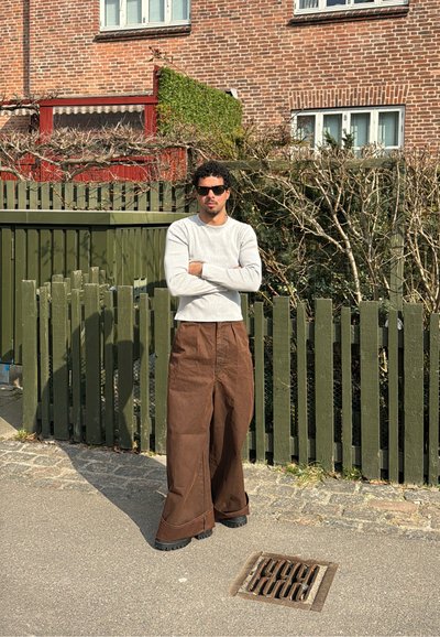 Camisa de manga larga de rayas en gris claro, pantalones anchos marrones, gafas de sol, de pie cerca de una cerca de madera verde y un fondo de pared de ladrillo.