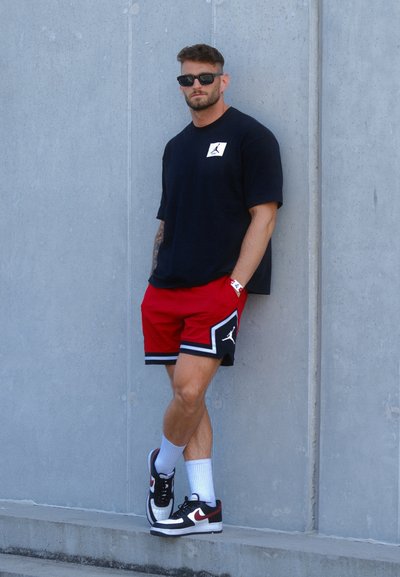 Camiseta negra de manga corta con logo, pantalones cortos rojos con detalles negros, calcetas blancas y zapatillas negras/rojas. Fondo de concreto.