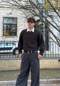 Vyras su garbanotais plaukais ir saulės akiniais, dėvintis tamsiai rudos spalvos megztinį ant balto marškinių su apykakle, tamsias platėjančias kelnes, stovintis lauke prie tvoros.