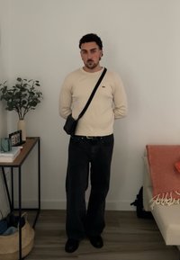 Cremfarbener, gerippter Langarmshirt, schwarze weit geschnittene Jeans und eine schwarze Umhängetasche. Inneneinstellung mit Pflanzen und minimaler Dekoration.