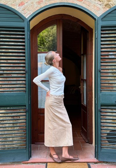 Falda maxi de denim beige claro con bolsillos laterales, combinada con una blusa ajustada de manga larga blanca de canalé y bailarinas marrones, de pie en una puerta.
