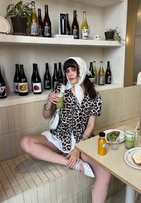 Camisa de manga curta com padrão de leopardo, saia branca, sapatos brancos com estampas. Bebida verde num copo, cercada por várias garrafas em prateleiras.