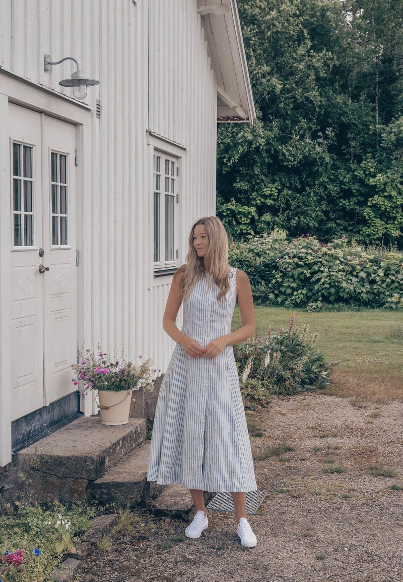 Jeune femme en robe sans manches rayée et baskets blanches debout près d'une maison en bois blanche avec des fleurs en pot sur les marches.