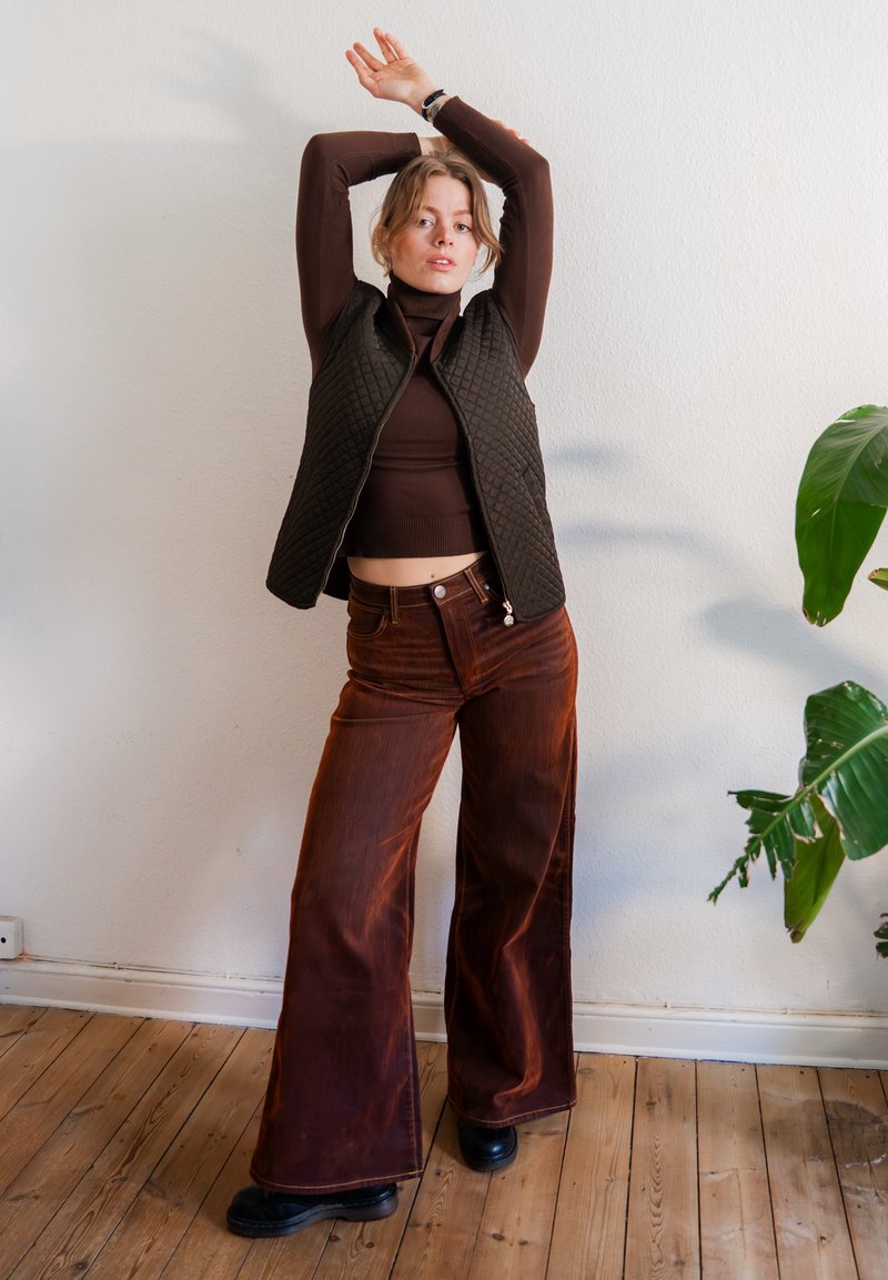 Person iført brun rullekrave, vatteret vest og bukser med vide ben strækker armene op over hovedet og står på trægulv ved siden af en grøn plante.