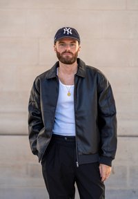 Μαύρο δερμάτινο τζάκετ μπομπέρ με φερμουάρ, φορεμένο πάνω από λευκό φανελάκι. Συνδυασμένο με μαύρο παντελόνι και ναυτικό καπέλο με λευκό λογότυπο.