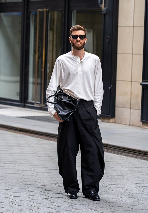 Mann mit Bart und Sonnenbrille geht die Straße entlang, trägt ein weißes langärmeliges Hemd, weite schwarze Hose, schwarze Schuhe und hält eine schwarze Tasche.