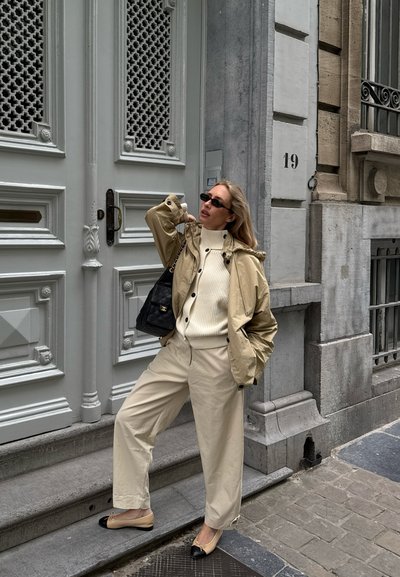 Mujer con conjunto beige y gafas de sol posando junto a una puerta blanca en una calle de la ciudad, llevando un bolso negro y calzando zapatos planos beige.