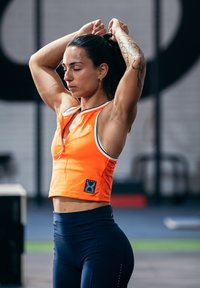 Oranžs sporta apgrieztais tops ar rāvējslēdzēju un baltām akcentēm, pārī ar augsto jostasvietu tumši zilām legingām. Topā ir logotipa uzšuvums.
