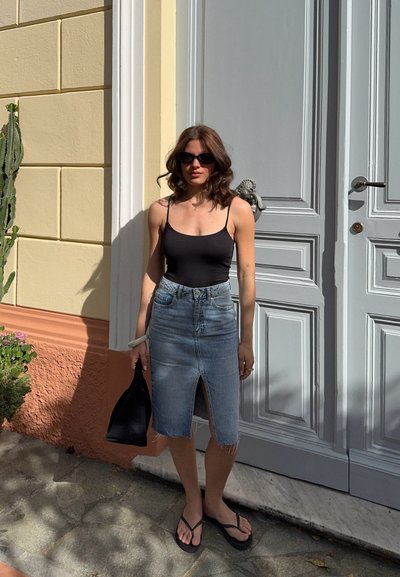 Mujer con gafas de sol, camiseta sin mangas negra, falda vaquera con abertura frontal y chanclas, está de pie sosteniendo una bolsa negra frente a una puerta gris.