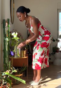 Květinové midi šaty s červenými a zelenými hibiskovými vzory, vyrobené z lehké tkaniny. Bílé vysoké podpatky, průhledná váza a zelené rostliny na dřevěném stole.