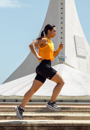 Atleta con una camiseta sin mangas de color naranja brillante y pantalones cortos negros, corriendo por escalones de concreto. Zapatillas de correr grises con recortes únicos visibles.