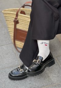 Μαύρα λουστρινένια loafers με χοντρές σόλες, φορεμένα με λευκές κάλτσες που έχουν ναυτικό κείμενο και μια κόκκινη καρδιά. Καλάθι από πλεκτό ξύλο στο φόντο.