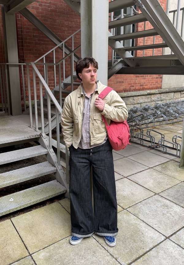 Unge med krøllet hår iført beige jakke, brede sorte bukser og blå sko, der holder en rød rygsæk nær metaltrapper og cykelstativer.