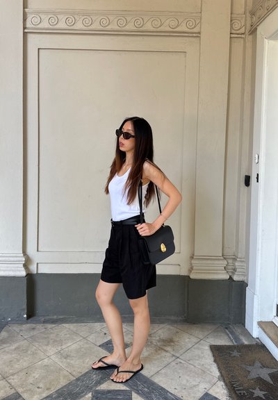 Pantalones cortos negros con pliegues, camiseta sin mangas blanca y chanclas negras. Lleva un bolso negro con un cierre dorado. Cabello largo y castaño, con gafas de sol puestas.