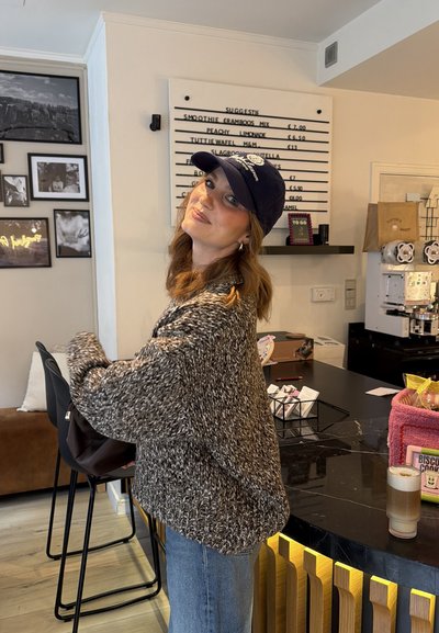Mujer con suéter de punto y gorra, de pie dentro de una cafetería cerca de un cartel de menú y un mostrador con aperitivos y una máquina de café.