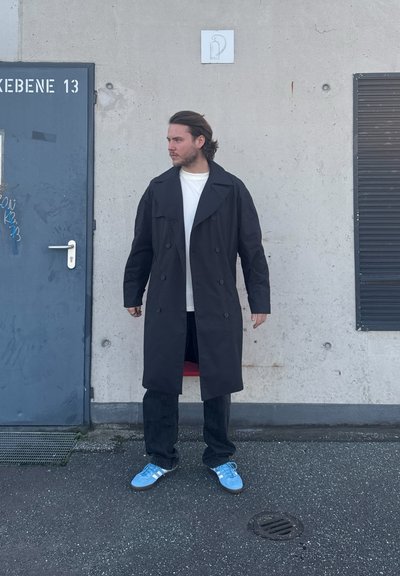 Gabardina negra de corte cruzado con solapas anchas, llevada sobre una camisa blanca, combinada con jeans negros y zapatillas azul claro. Fondo de pared de concreto.