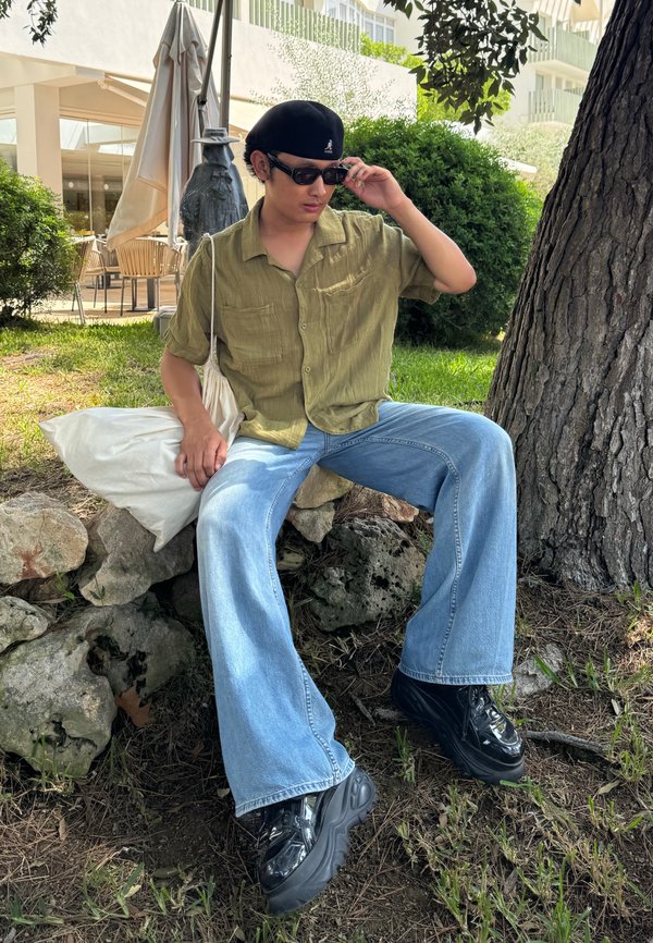 Chemise verte à manches courtes, jean large bleu clair et chaussures à plateforme noires. Sac blanc reposant sur une surface en pierre près d'un arbre.