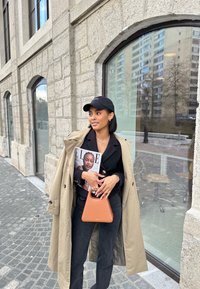 Beige trenchcoat över en svart topp, hållande en tidning och en orange handväska. Svart keps, mörka byxor, utomhusmiljö med sten och glas.