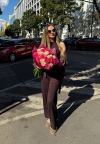 Bukiet różowych i kremowych róż owinięty w różowy papier, trzymany przez osobę w ciemnobrązowym stroju i okularach przeciwsłonecznych, stojącą na ulicy miasta.
