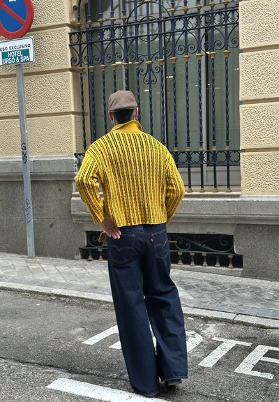 Suéter de punto a rayas amarillas y negras, diseño recortado, combinado con jeans oscuros y holgados. Gorra casual y postura relajada en un entorno urbano.