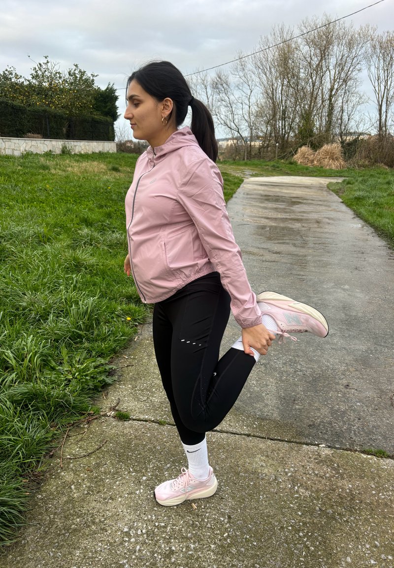 Žena v ružovej bunde a čiernych legínach natiahuje jednu nohu na ceste vedľa trávy a holých stromov pod zamračenou oblohou.
