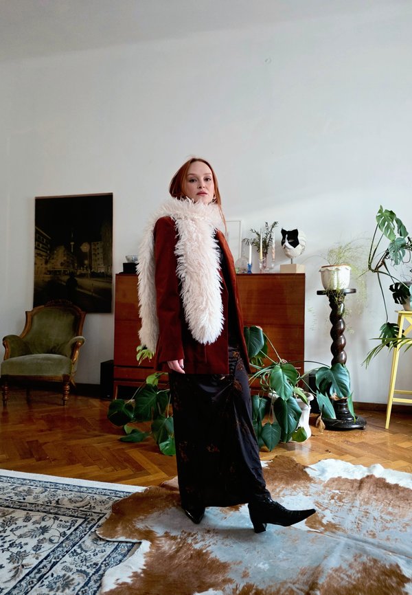 Ženska v dolgi temni krilu, rjavi jakni in puhastem belem šalu stoji v sobi z rastlinami, starinskim stolom in lesenim tlemi.