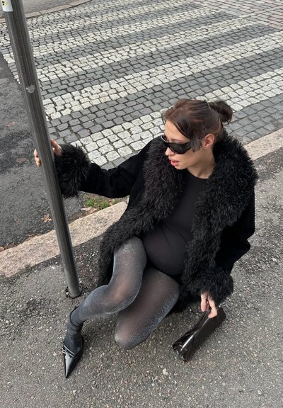 Mujer con abrigo de piel negro y medias brillantes sentada en la acera cerca de un paso de cebra, sosteniendo un poste con una mano y un bolso con la otra.