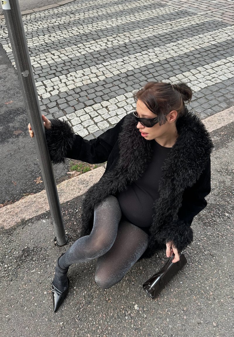 Femme portant un manteau en fourrure noire et des collants scintillants assise sur le trottoir près d'un passage pour piétons, tenant un poteau d'une main et un sac à main de l'autre.