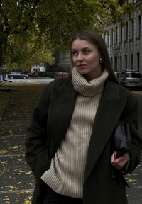 Μοντέλο που φοράει ένα σκούρο πράσινο μάλλινο παλτό πάνω από ένα μπεζ ribbed πουλόβερ, κρατώντας μια μαύρη τσάντα-φάκελο. Δρόμος με φθινοπωρινά φύλλα και αυτοκίνητα στο παρασκήνιο.