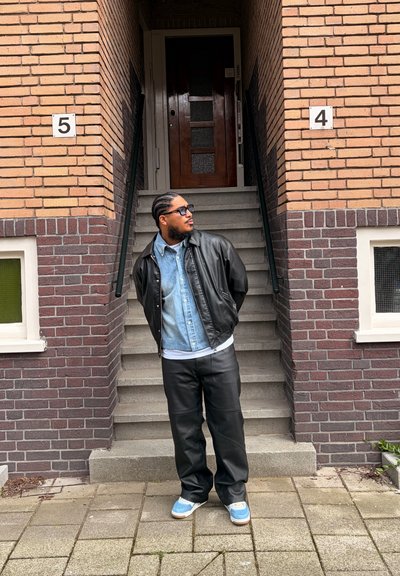Hombre con el pelo trenzado, gafas de sol, chaqueta de cuero, camisa vaquera y zapatillas, de pie frente a los escalones de un edificio de ladrillo numerados 4 y 5.