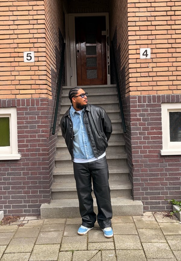 Mann mit geflochtenen Haaren, Sonnenbrille, Lederjacke, Jeanshemd und Turnschuhen steht vor den Backsteintreppen mit den Nummern 4 und 5.