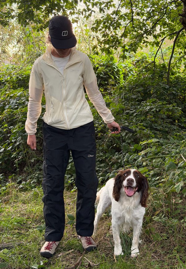 Persona con ropa de exterior paseando a un perro blanco y marrón alegre con correa en una zona verde y arbolada.