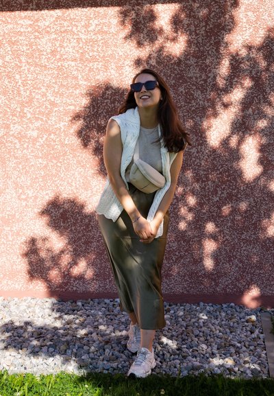 Chaleco de punto blanco, blusa beige, falda de satén verde oliva, bolso bandolera de crema y zapatillas blancas contra una pared texturizada rosa con piedras abajo.