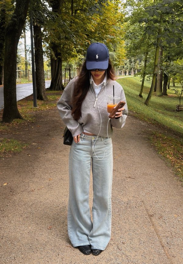 Femme portant un sweat-shirt gris et une casquette bleue se tient sur un chemin bordé d'arbres tenant un téléphone et une boisson glacée avec des écouteurs dans un parc.
