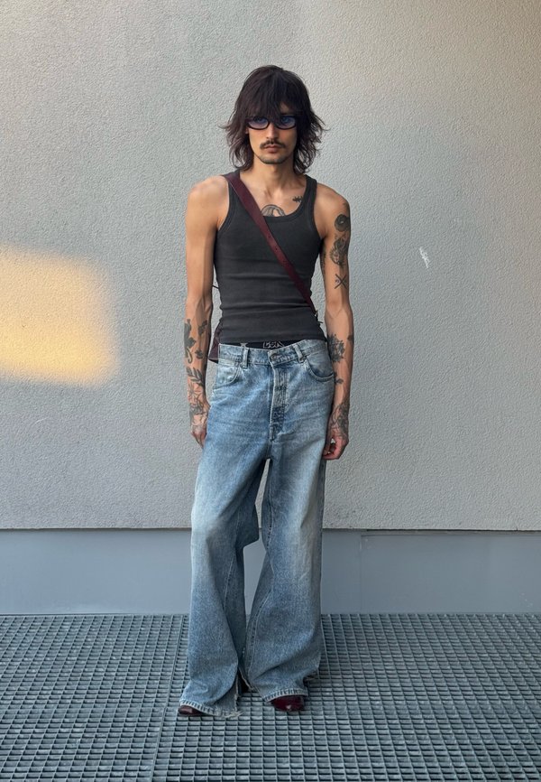 Personne mince aux cheveux mi-longs, portant des lunettes de soleil, un débardeur noir, un jean large délavé et un sac bandoulière, debout contre un mur gris.
