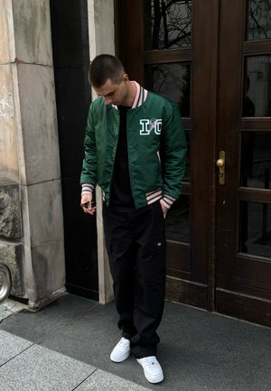 Chaqueta bomber de satén verde con un cuello y puños de canalé rosa, presenta un diseño gráfico. Combinada con pantalones cargo negros y zapatillas blancas.