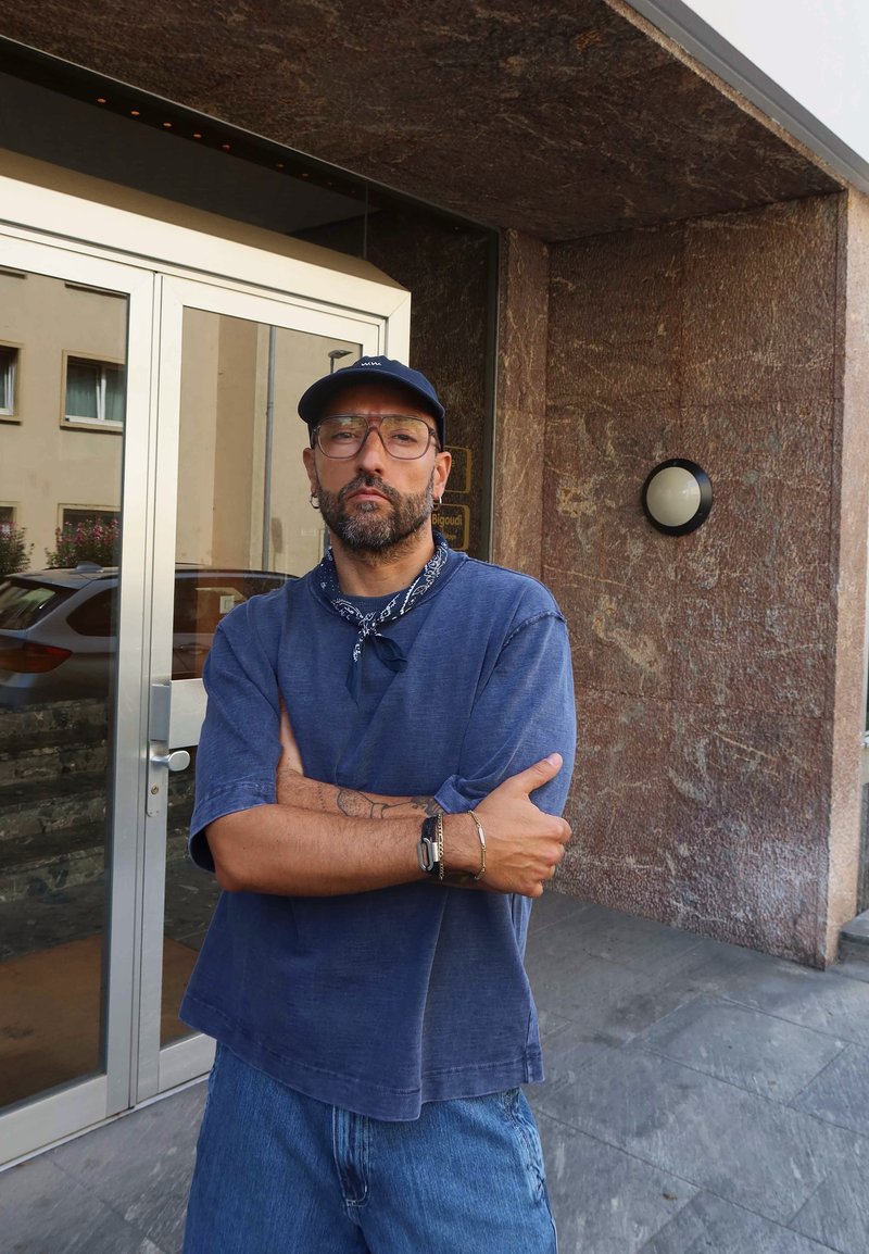 Uomo con barba e occhiali che indossa un cappello, una camicia blu, pantaloni corti di jeans e un bandana attorno al collo, in piedi con le braccia incrociate all'esterno dell'ingresso di un edificio.