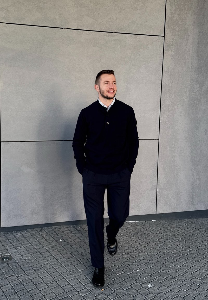 Young man in black jumper and trousers walking on tiled ground with hands in pockets against grey concrete wall, smiling and looking right.