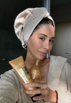 Two Redken haircare products: a golden conditioner and a golden tube of leave-in treatment. The person wears a beige towel on their head and a textured robe.