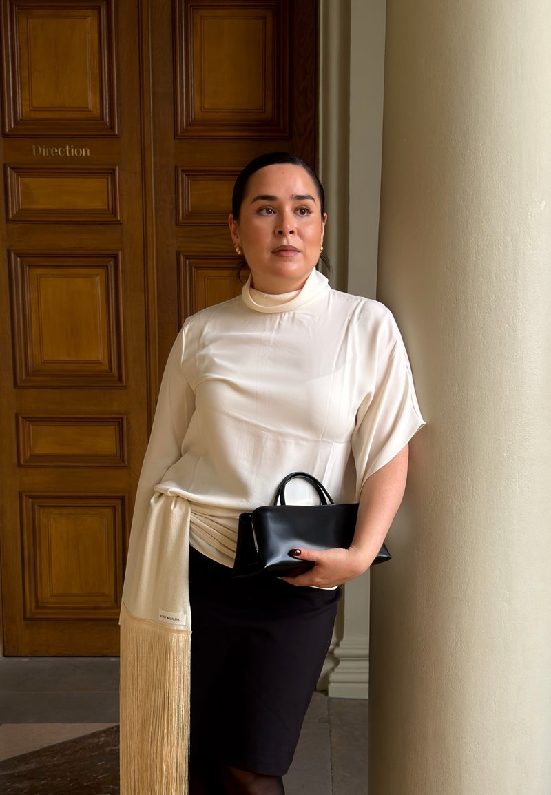 Weiße Bluse mit hohem Kragen, drapiertem Design und Fransen-Detail; schwarze, taillierte Rock; trägt eine kleine schwarze Handtasche. Holztür als Hintergrund.