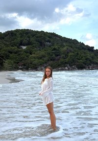 Copricostume in lino bianco con maniche lunghe e orlo con coulisse, in piedi in acqua bassa sullo sfondo di colline verdi e cielo nuvoloso.