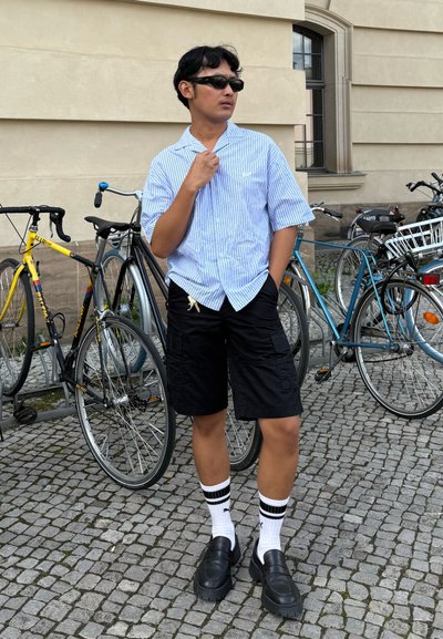 Camisa de rayas azul y blanca de manga corta, pantalones cortos de carga negros, zapatos de plataforma negros y calcetines blancos hasta la rodilla con rayas negras, de pie junto a bicicletas.