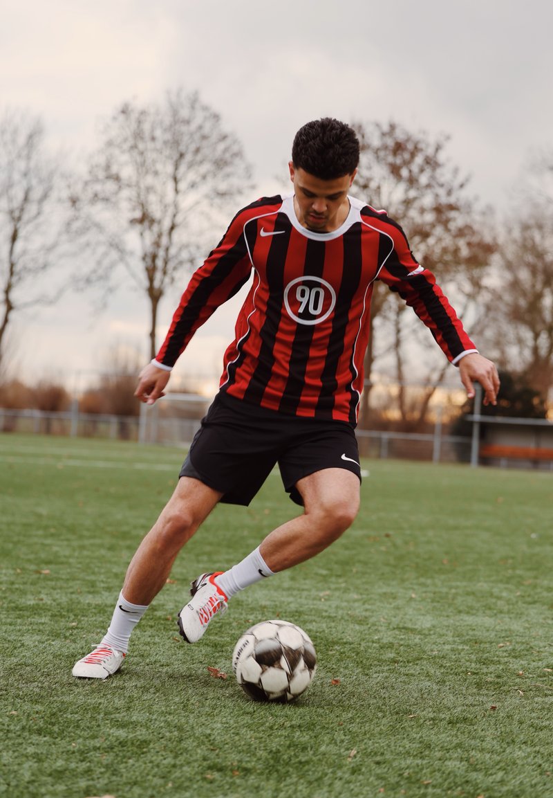 Jeune homme portant un maillot rayé rouge et noir avec le numéro 90, un short noir et des crampons blancs contrôlant un ballon de football sur un terrain herbeux.